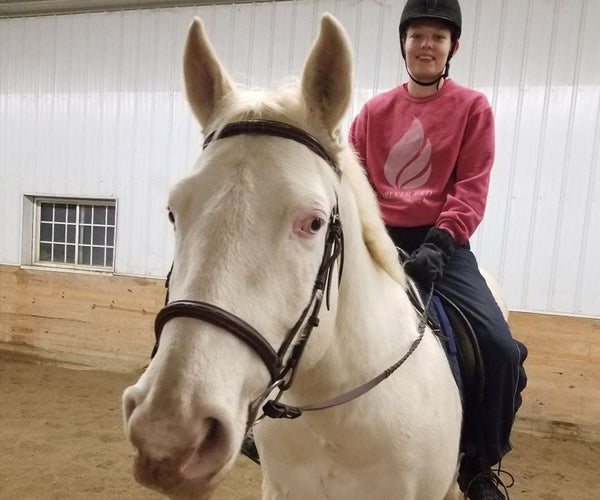 A woman in a pink jumper and black helmet riding a cream coloured horse with a black bridle in a white and yellow sand arena